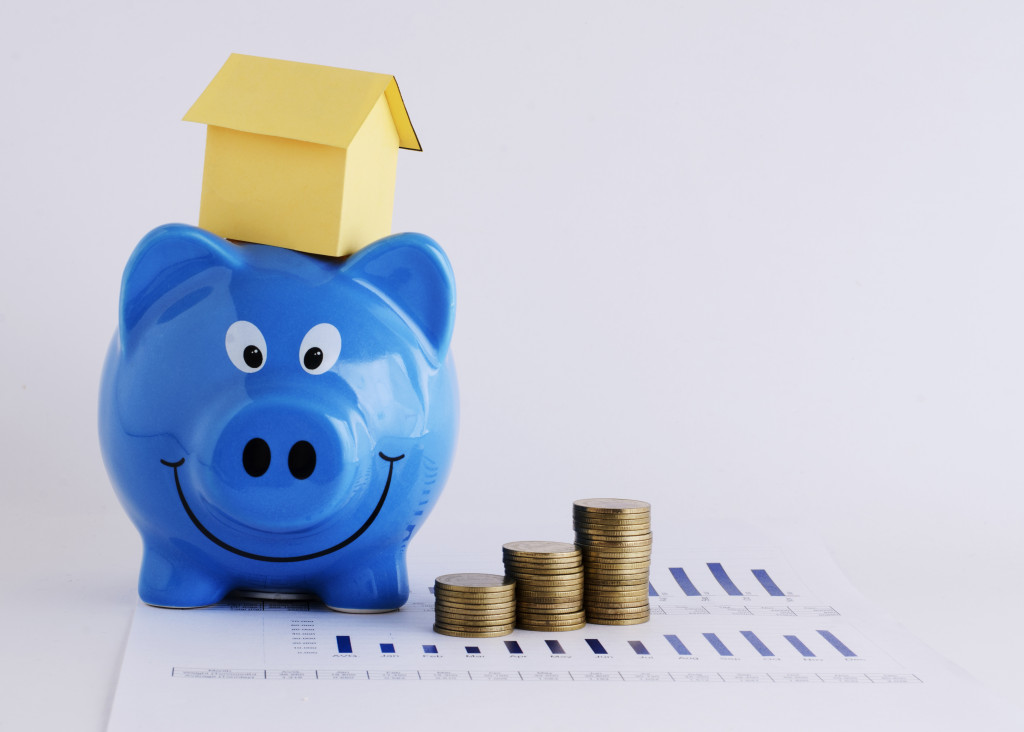 A miniature house on a blue piggy bank with stacks of coins beside it